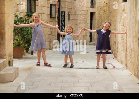Drei junge Schwestern/Mädchen/Kinder/Kinder/Kind 7, 3, & 5 Jahre, für einen Familienurlaub, laufen und spielen und über eine schmale Straße in Mdina, Malta Stockfoto