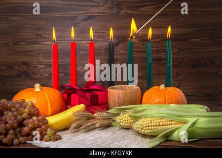 Zündung von kwanzaa traditionelle Kerzen, festival Konzept mit Geschenkbox, Kürbisse, Ohren von Weizen, Trauben, Hühneraugen, Banane, Schüssel und Früchte auf Holz- Bac Stockfoto