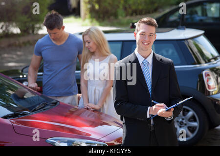 Junge Verkäufer schriftlich über die Zwischenablage mit ein paar auf der Suche nach neuen Auto im Hintergrund Stockfoto