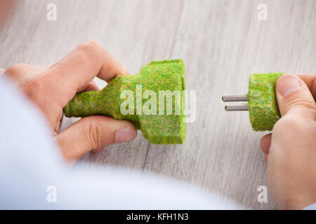 Zugeschnittenes Bild der Geschäftsmann mit Rasen bedeckt, elektrische Stecker und Steckdose am Schreibtisch im Büro Stockfoto