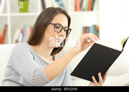 Porträt einer Frau, die das Tragen von Brillen lesen Sie ein Buch aus Papier auf einem Sofa zu Hause sitzen Stockfoto