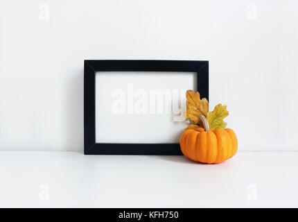 Schwarze leere Holzrahmen mockup mit einem orangefarbenen Kürbis und Golden Oak Blätter liegen auf dem weißen Tisch. Plakat Produktdesign. styled lieferbar feminine Fotografie. Wohnkultur. Herbst, Herbst Konzept. Stockfoto