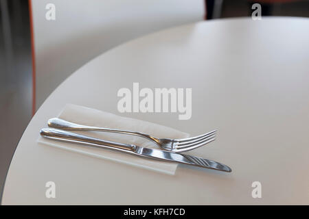 Silbermesser und Gabel auf weißer Papierserviette auf dem weißen runden Tisch mit verschwommener Stuhlrückseite im Hintergrund Stockfoto