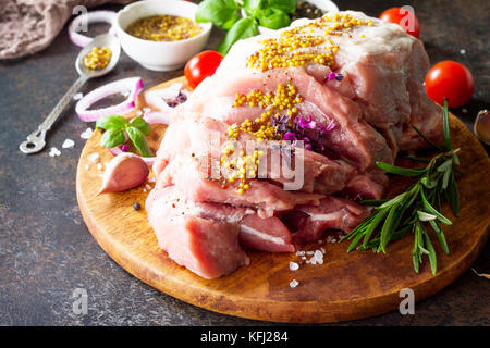 Schweinefilet mit Senf mariniert auf einem Schneidebrett, rohes Fleisch, Tomaten und verschiedene Gewürze auf einem Stein oder Schiefer Hintergrund. Stockfoto