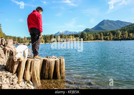 Eine asiatische Junge in rot Fleece steht auf einem Baumstumpf mit seinen weißen Hündchen am Rande eines Sees in einem Herbst sonnigen Nachmittag Stockfoto