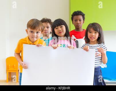 Vielfalt Kinder Holding leeres Plakat im Klassenzimmer an Kindergarten Vorschule, multiethnische Gruppe mit Schild, Mock up für Hinzufügen von Text oder Design. Stockfoto