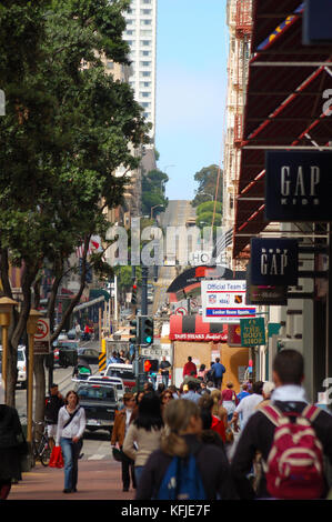 Besetzt Käufer an der Unterseite der Powell Street in San Francisco. Menschen und Schilder im Vordergrund, von Bäumen eingerahmt auf der linken Seite, Seilbahnen in der Nähe von der Spitze des Hügels im Hintergrund, vor blauem Himmel. Stockfoto