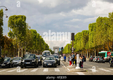Autos gestoppt an der Ampel, auf den von Bäumen gesäumten Champs-Elysee in Paris, Frankreich, Menschen warten auf einen Zebrastreifen überqueren, der Triumphbogen im Hintergrund unter einem blauen bewölkten Himmel. Kopieren Sie Platz. Stockfoto