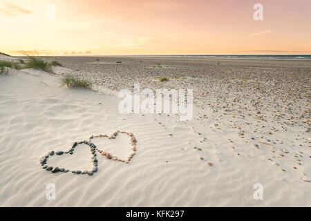 Pebbles angeordnet in Form von zwei Herzen auf Sand strand Wellen mit schönen Sonnenuntergang. Stockfoto