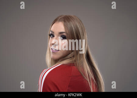 Junge blonde Frau in retro rot Trainingsanzug zurück über die Schulter auf der Kamera über Grau studio Hintergrund drehen. Stockfoto