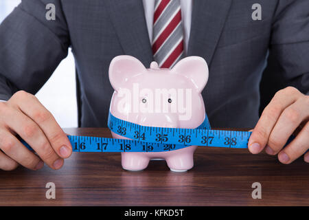 Mittelteil der Geschäftsmann messen Sparschwein mit Maßband am Schreibtisch Stockfoto