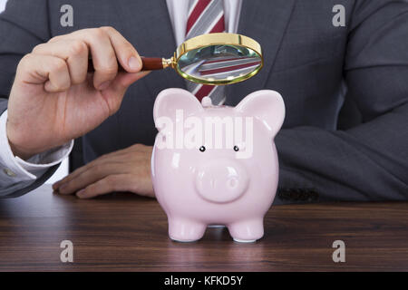 Mittelteil der Geschäftsmann Prüfung Sparschwein mit Lupe am Schreibtisch Stockfoto