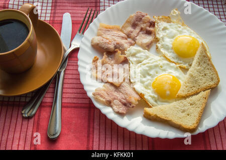 Köstliches Frühstück - eine Tasse Kaffee, einen Teller mit Spiegelei, Speck und Toast, neben dem Besteck auf rot kariert Serviette. Stockfoto