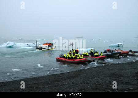 Touristen in Tierkreisbooten, die das Ufer verlassen, auf der Jokulsarlon Gletscherlagune, die Eisberge in leuchtend gelben Lebenswesten, Südisland, betrachtet Stockfoto