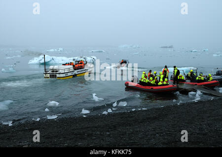 Touristen in Tierkreisbooten, die das Ufer verlassen, auf der Jokulsarlon Gletscherlagune, die Eisberge in leuchtend gelben Lebenswesten, Südisland, betrachtet Stockfoto