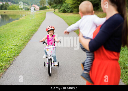 Süße kleine glückliche Mädchen mit ihrer Mutter und ihrem jüngeren Bruder Kleinkind in der Natur am See. Mädchen ist Radfahren und Smilling. slow motion. Stockfoto