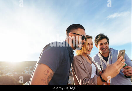 Gruppe der tausendjährigen über ein Mobiltelefon an Party im Freien. Lächelnde junge Männer und eine Frau stehen auf dem Dach und mit Handy. Stockfoto