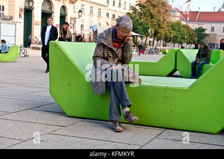 Ältere kaukasische Frau sitzt auf einer modernen grünen Bank am Museumsplatz, spricht auf dem Handy. Rentner im höheren Alter. Wien, Österreich, Europa EU Stockfoto