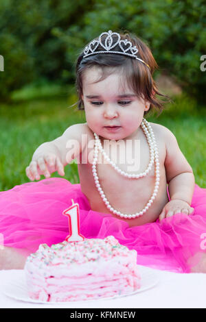 Smiling Happy Baby Kleinkind Mädchen in der ersten Geburtstagsparty mit rosa Kuchen outdoor. gekleidet mit Princess Tiara, Perlenkette, Tutu für Jahrestag Stockfoto