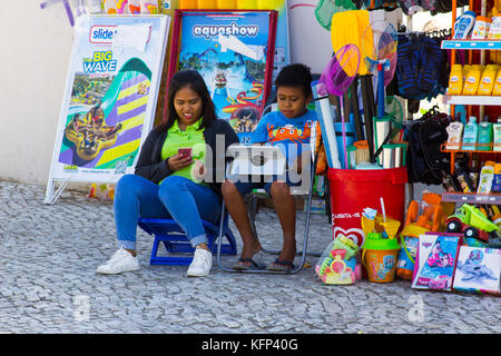 Mutter und Sohn benutzen ihr Handy und Tablet, während sie vor ihrem kleinen Laden in Albuferia in Portuga sitzen Stockfoto