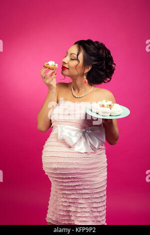 Porträt eines hungrigen glücklich Schwangere Frau essen eine süße Kuchen, auf einem rosa Hintergrund isoliert. Stockfoto
