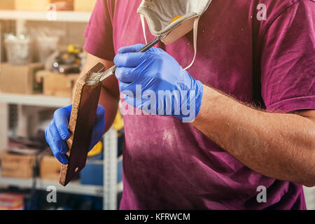 Eine Nahaufnahme eines männlichen Tischler in ein Lila T-Shirt und blaue Handschuhe sanft malt eine Holz- Quaste mit einem Holzblock in eine dunkelbraune Farbe Stockfoto