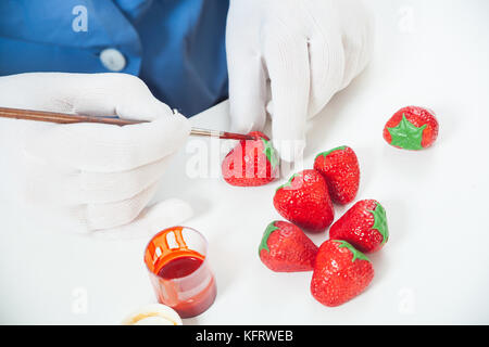 Eine Frau Konditor mit blauen Uniform und weiße, sterile Handschuhe, malt ein nophet in Form einer Erdbeere in Rot mit essbarer Farbe auf einem weißen Tisch Stockfoto