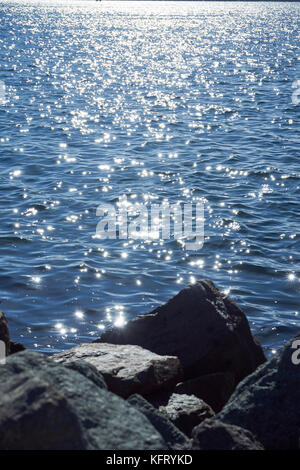 Steine und Felsen im Vordergrund der glitzernden Sonne auf dem Wasser Stockfoto