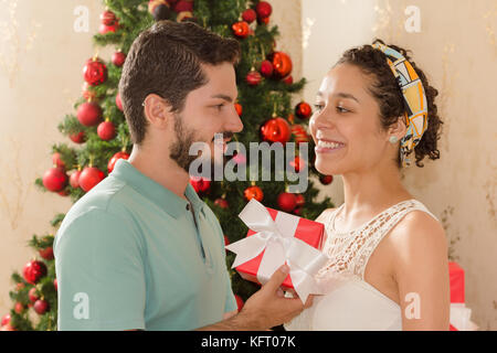 Freund ist die Übergabe der Weihnachtsgeschenk für eine Frau. Freundin ist glücklich und lächelnd. Weihnachtsbaum im Hintergrund eingerichtet. roter Geschenkbox Stockfoto