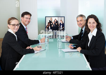 Gruppe von männlichen und weiblichen Geschäftsleuten an einem Tisch sitzt gerade eine online Präsentation auf einem Bildschirm Stockfoto