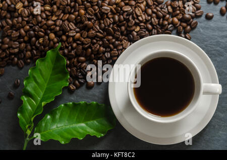 Kaffeetasse und geröstete Kaffeebohnen mit grünen Blättern auf Schwarz, dunklen steinernen Tisch Hintergrund Stockfoto