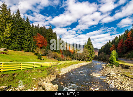 Kleiner Fluss in Fichten bewaldete Berge mit ein paar Bäumen in rot Laub. Campingplatz hinter dem Zaun auf einer grünen Wiese. wunderschöne Landschaft unter v Stockfoto