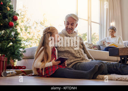 Ältere Menschen und Enkelin mit digitalen Tablette zu Hause während Weihnachten, mit älteren Frau auf Sofa im Rücken sitzen. Stockfoto