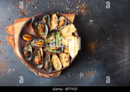 Overhead von Seafood Dinner Table. Gegrillte gebackene Muscheln mit Butter, Petersilie und Käse auf Keramikplatte auf dunklem Hintergrund. Ansicht von oben. Platz kopieren Stockfoto