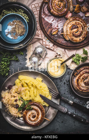 Gebratene Würstchen mit Kartoffelbrei und Sauerkraut auf dunklen rustikalen Tisch mit Senf serviert, Ansicht von oben. Die Deutschen traditionelles Essen Stockfoto