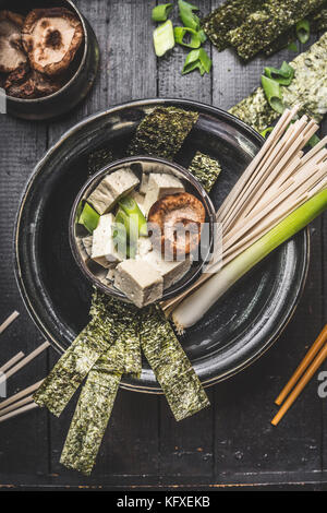 Asiatische Suppe Vorbereitung in Schüssel mit udon noodls, Tofu, Shiitake-Pilze, Nori und Frühlingszwiebeln auf dunklen rustikalen Hintergrund, Ansicht von oben Stockfoto