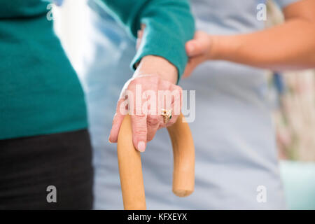 Die ältere Frau Hände auf Gehstock mit Sorgfalt Arbeiter im Hintergrund Stockfoto