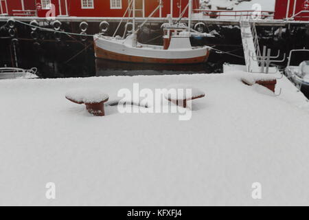 Schnee bedeckt Hafen von nusfjord Fischerdorf mit schwimmenden Pontons und Fischerboote in - rostige Poller - rot Holz- fischerhütten oder Rorbuer fo günstig Stockfoto