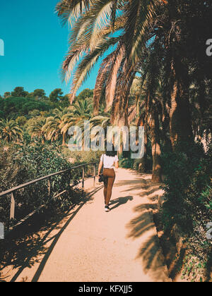 Junge trendige Schöne gebräunte Frau mit schwarzen Haaren Spaziergänge entlang Pfad zwischen tropischen Palmen in Barcelona Park Güell. travel Concept. Stockfoto