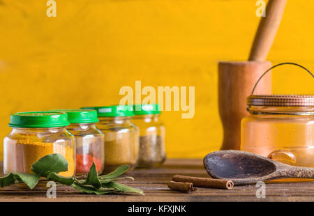 Gewürze Container, Töpfe und Schalen aus Glas in einer Reihe, auf einer hölzernen Oberfläche, mit Mörser und Stößel, Lorbeerblatt, Holzlöffel und Zimt Dekor Stockfoto