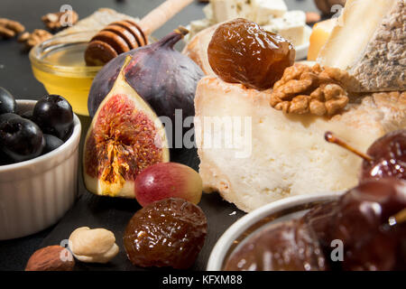 Die Zusammensetzung von Lebensmitteln mit Stück Käse, Trauben, Datteln, Oliven, Nüsse, Honig, Feigen auf dunklem Hintergrund. Stockfoto