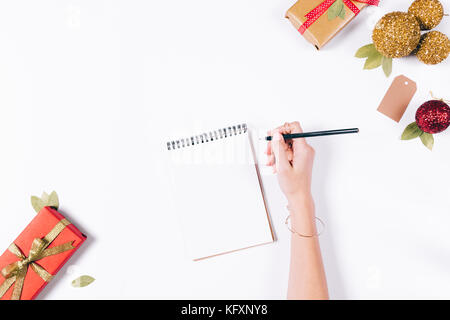 Weibliche Hand schreibt in einem Notebook mit einem Bleistift auf einem weißen Tisch unter Geschenke des neuen Jahres, eine Ansicht von oben Stockfoto