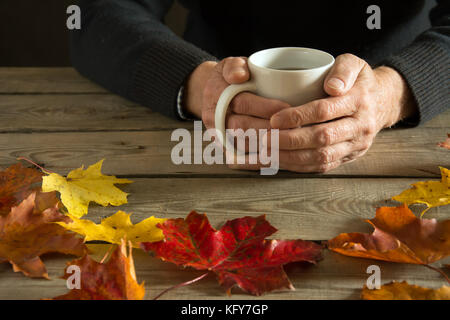 Die Hände eines alten Mannes halten einer Tasse auf einem Holztisch mit bunten Blättern Stockfoto