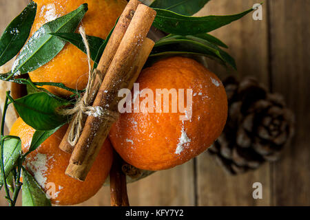 Lebhaften orange Mandarinen auf den Ästen grüne Blätter Cinnamon Sticks mit Garn gebunden Pine Cone Holz Hintergrund. Weihnachten Neujahr Grußkarten Poster Stockfoto