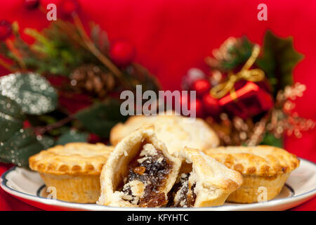 Nahaufnahme eines gebrochenen Mince Pie auf einer Platte mit einigen festlichen Weihnachtsschmuck auf rotem Hintergrund Stockfoto