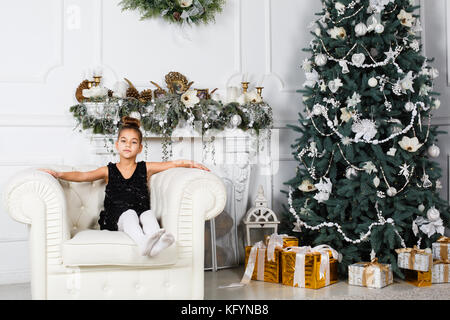 Kleines Mädchen sitzt auf einem Sessel im Zimmer mit Weihnachtsbaum und Geschenkboxen auf dem Boden Stockfoto