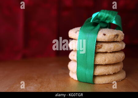 Stapel von fünf Choc Chip und Obst Plätzchen, in einem grünen Band umwickelt, auf einem Holztisch mit einem festlichen roten Hintergrund. Kopieren Sie Platz für Text.. Kopieren spac Stockfoto