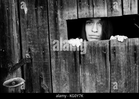 Junge Frau auf der Suche Durch den Riss eines gesperrten Holzschuppen. schwarz-weiß Foto. Stockfoto