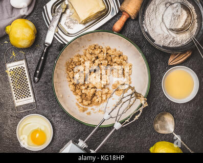 Zitrone Plätzchen oder Kuchen Zubereitung mit Zutaten zum Kochen. Butter und Zucker mischen mit der Hand Mixer auf dunklen Küche Tisch Hintergrund, Ansicht von oben Stockfoto