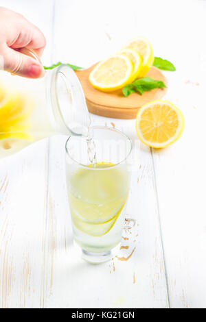 In einem Becherglas, eine kalte Limonade wird aus einer Kanne auf einem weißen Hintergrund Holz umgeben von Zitronen gegossen Stockfoto